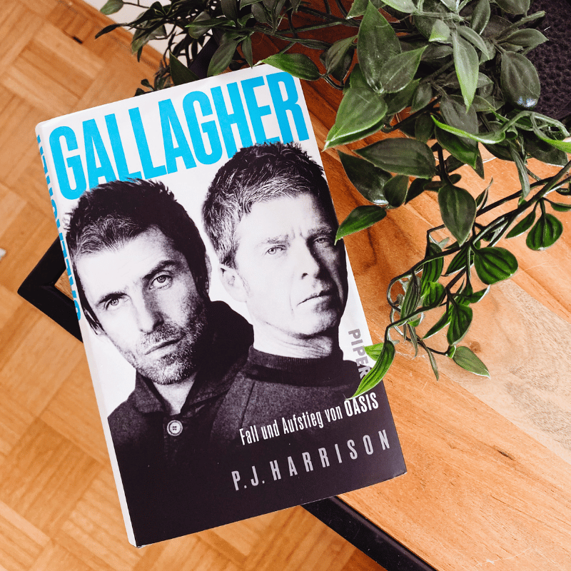 „Gallagher – Fall und Aufstieg von Oasis“ von P.J. Harrison liegt auf einem Tisch neben einer Pflanze