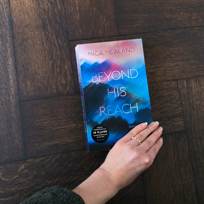 „Beyond his reach“ liegt auf einem dunklen Holzboden. Eine Hand liegt am Buch.