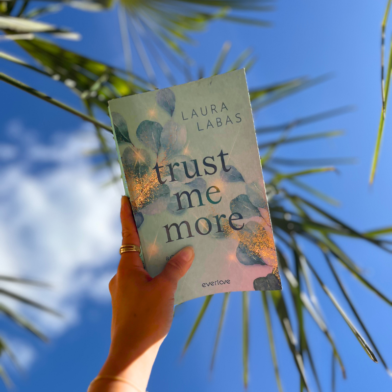 „Trust Me More“ von Laura Labas wird vor blauem Himmel mit Palmenblättern gehalten