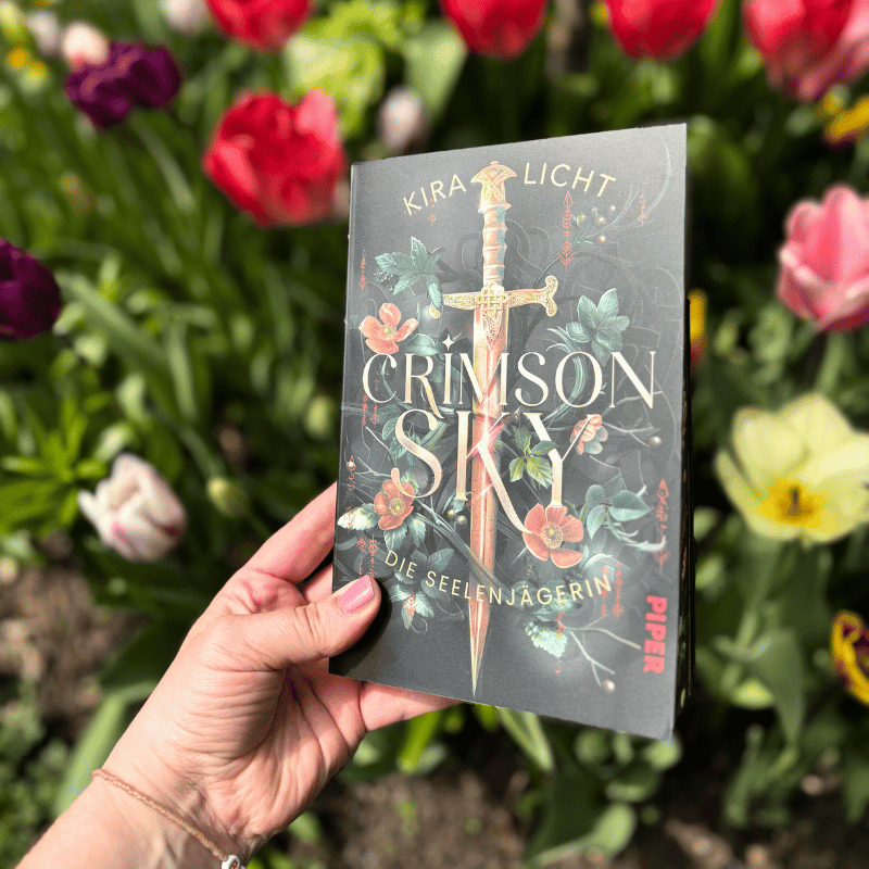 „Crimson Sky – Die Seelenjägerin“ von Kira Licht wird vor Blumen gehalten