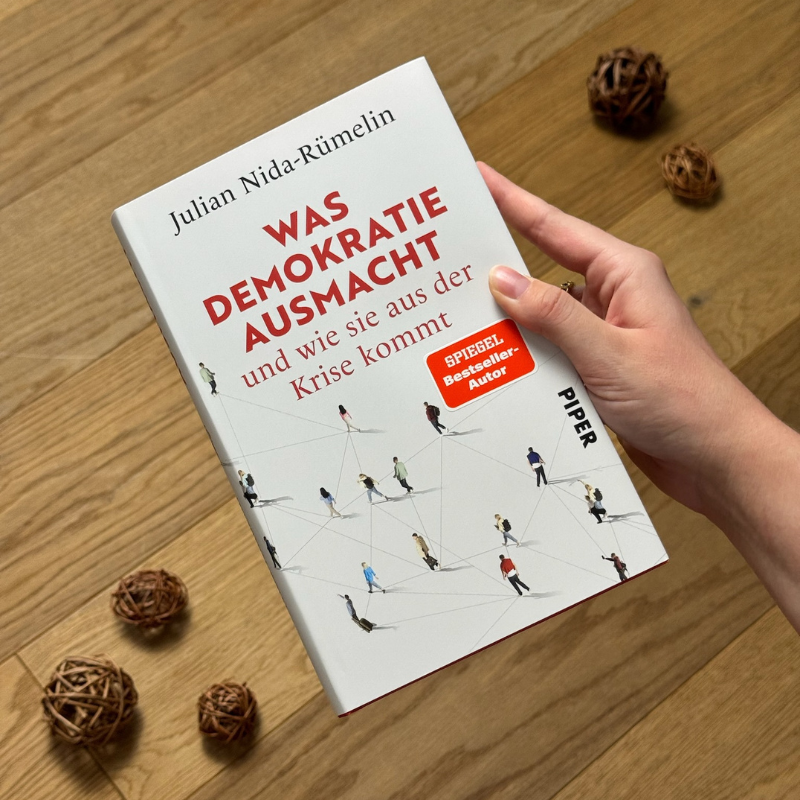 „Was Demokratie ausmacht“ von Julian Nida-Rümelin in einer Hand über einem Holzboden gehalten.
