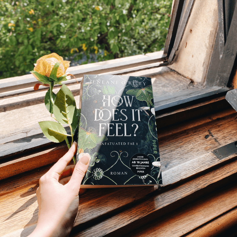 Eine Hand hält „How Does It Feel?“ von Jeneane O'Riley vor einem offenen Fenster.