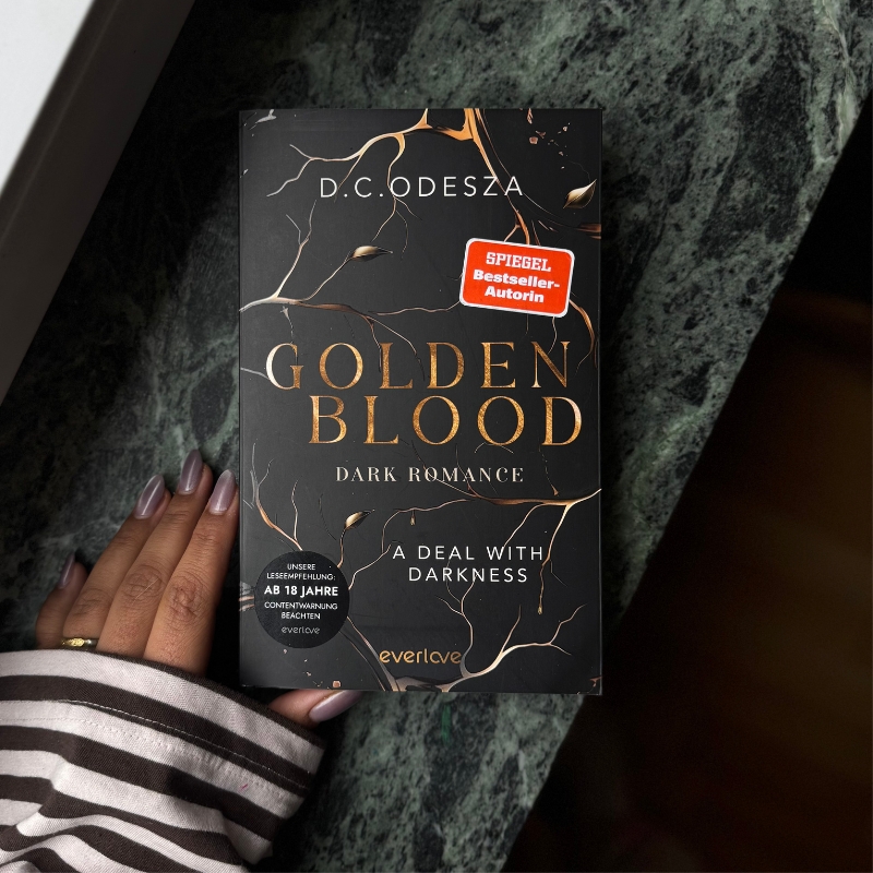 Eine Hand hält „Golden Blood – A Deal with Darkness“ auf einem dunklen Steinsims.