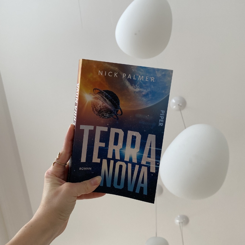 "Terra Nova" wird von einer Hand noch oben gehalten. Im Hintergrund ist die weiße Decke zu sehen, von der zwei weiße Kugellampen hängen.