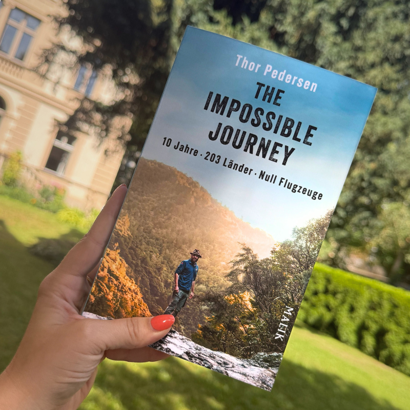 „The Impossible Journey“ von Thor Pedersen wird von einer Hand in die Luft gehalten. Im Hintergrund ist unscharf ein Garten zu erkennen.