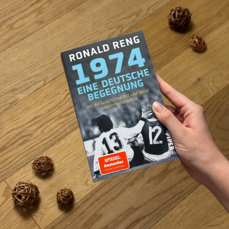 „1974 – Eine deutsche Begegnung“ von Ronald Reng in einer Hand über einem Holzboden gehalten.