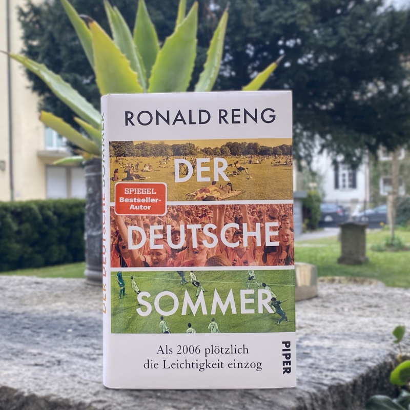 "Der deutsche Sommer" steht auf einer Mauer. Im Hintergrund ist ein Garten zu sehen.