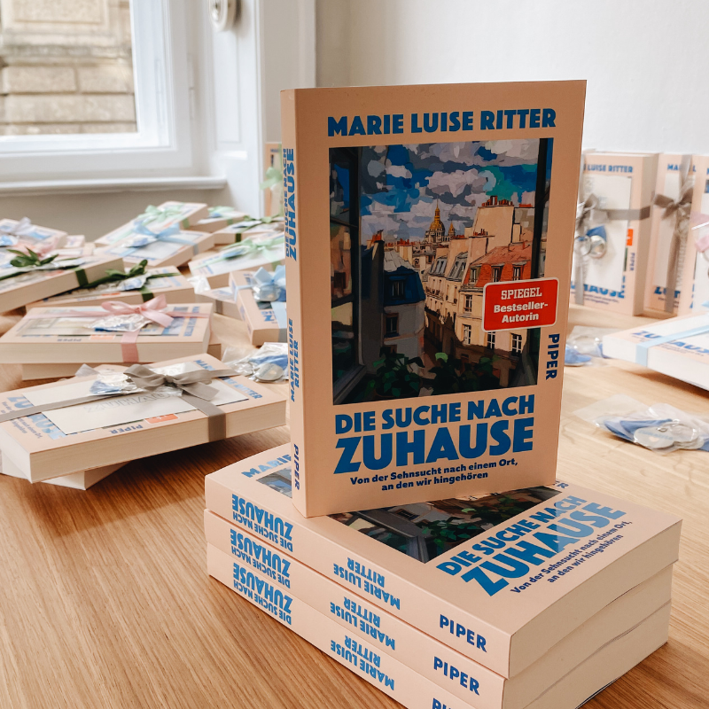 Mehrere Exemplare des Buches „Die Suche nach Zuhause“ von Marie Luise Ritter liegen gestapelt auf einem Tisch. Im Hintergrund sind weitere Bücher und Geschenke zu sehen.