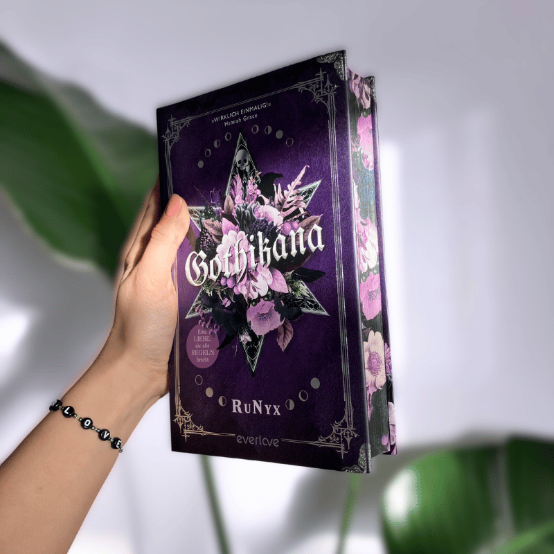 „Gothikana“ von RuNyx wird in einer Hand vor einer weißen Wand und einer Pflanze gehalten