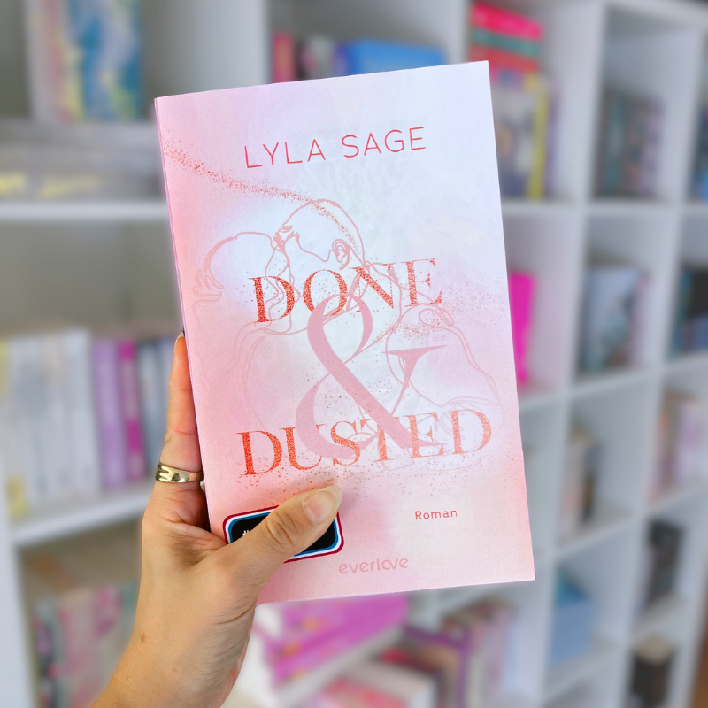 „Done & Dusted“ von Lyla Sage wird vor einem Bücherregal gehalten