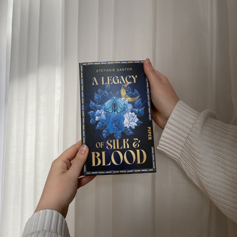 Zwei Hände halten "A Legacy of Silk and Blood" vor einem weißen Vorhang