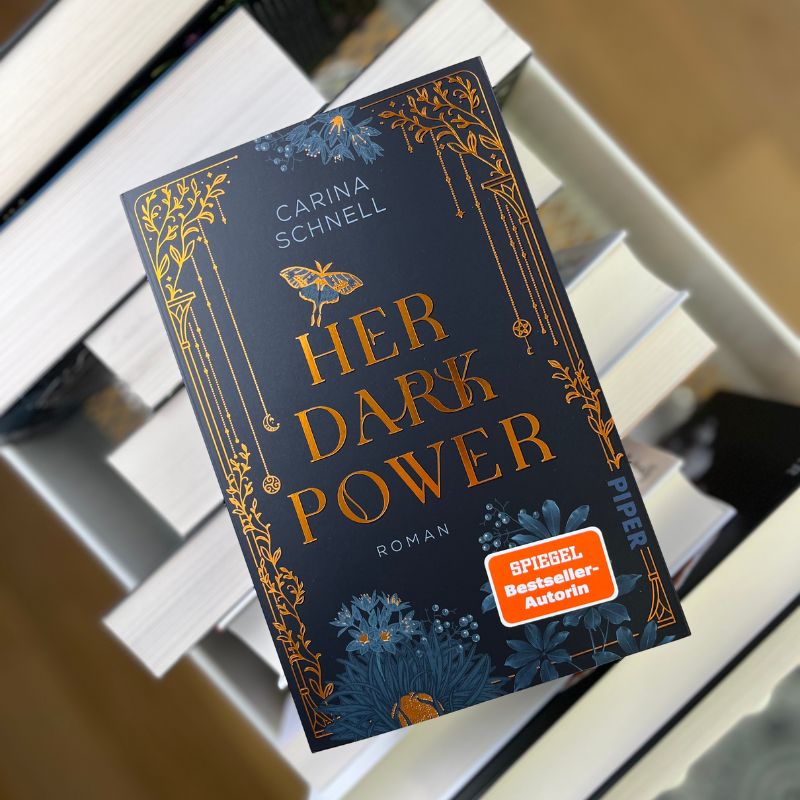 "Her Dark Power" von Carina Schnell liegt auf einem Bücherstapel.
