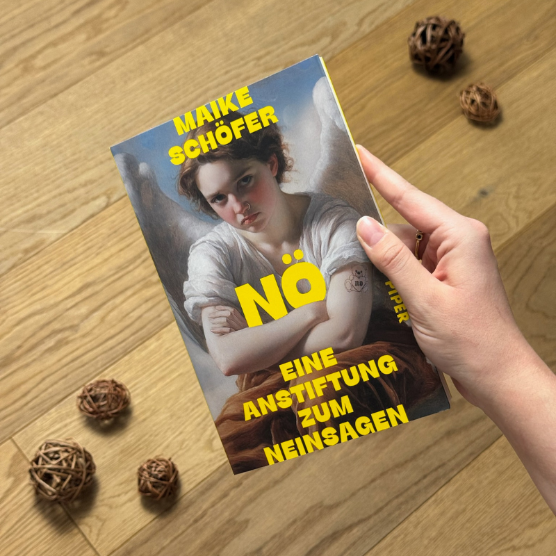 „Nö“ von Maike Schöfer in einer Hand über einem Holzboden gehalten.