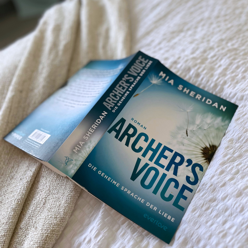 „Archer’s Voice“ von Mia Sheridan liegt aufgeschlagen mit dem Rücken nach oben auf einer weißen Decke.