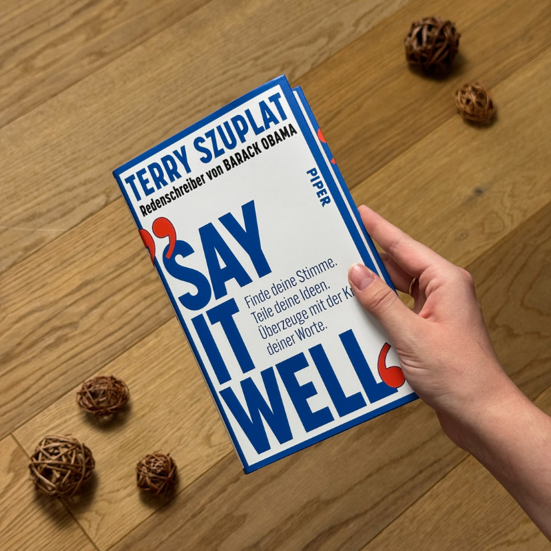 „Say It Well“ von Terry Szuplat in einer Hand über einem Holzboden gehalten.