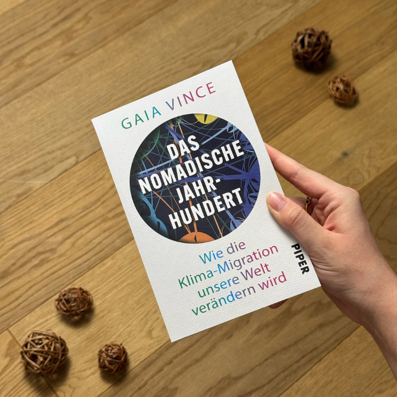 „Das nomadische Jahrhundert“ von Gaia Vince in einer Hand über einem Holzboden gehalten.