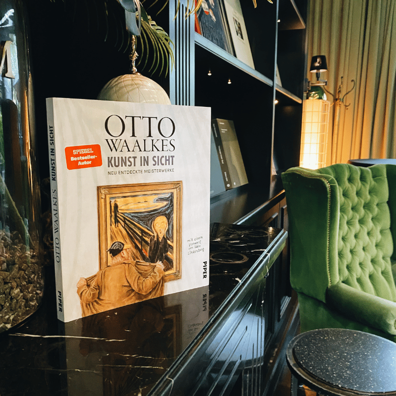 „Kunst in Sicht“ von Otto Waalkes steht auf einer dunklen Kommode, im Hintergrund ein schickes Regal und ein grüner Sessel