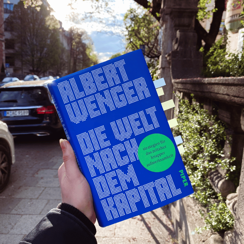 „Die Welt nach dem Kapital“ von Albert Wenger wird von einer Hand gehalten, im Hintergrund ist unscharf eine Straße zu sehen