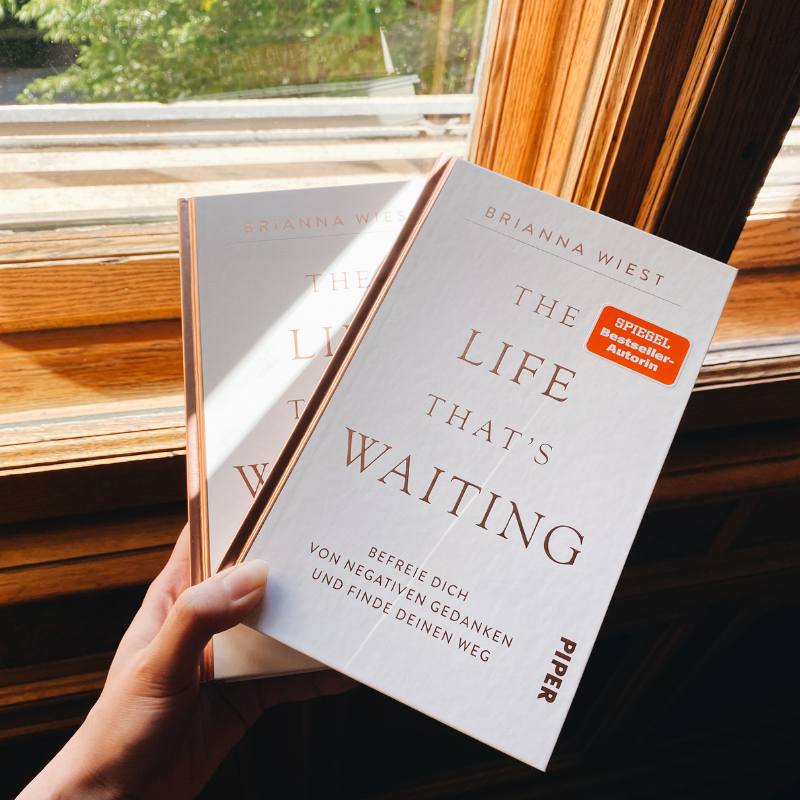 Eine Hand hält zwei Exemplare des Buchs „The Life That’s Waiting“ von Brianna Wiest vor einem geöffneten Holzfenster.