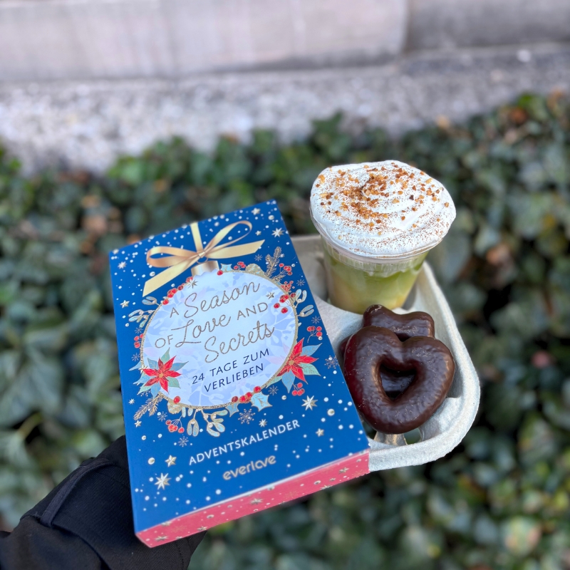 „A Season of Love and Secrets“ liegt neben einem Matcha Latte und Lebkuchenherzen auf einem To-Go-Kaffeehalter